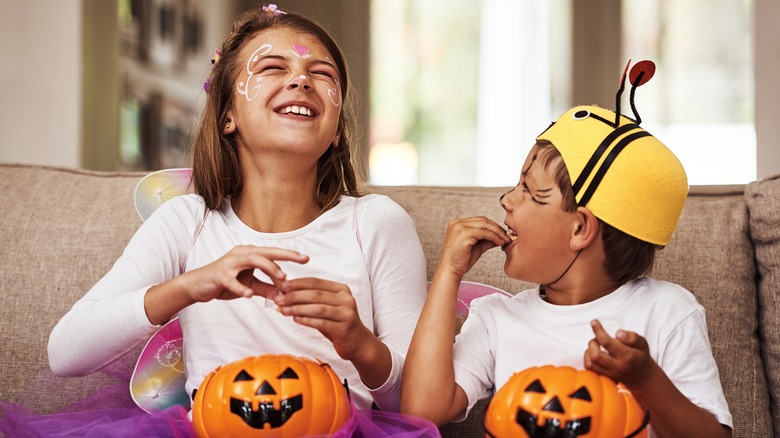 Two children sitting on the couch eating Halloween candy from plastic pumpkins