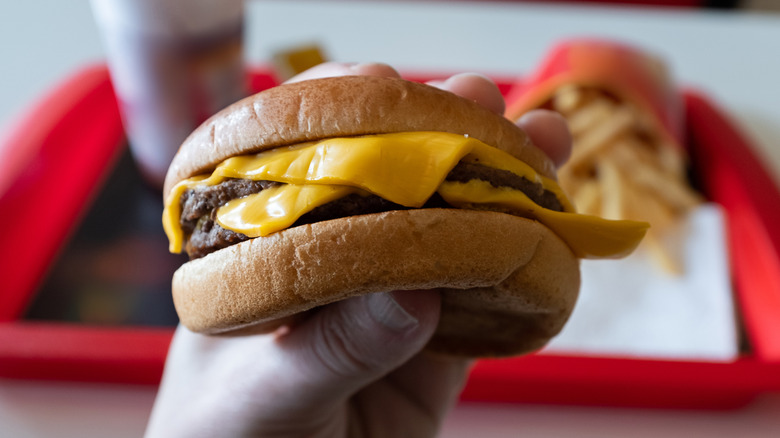 A man's hand holding a cheeseburger with a soda and fries in the background