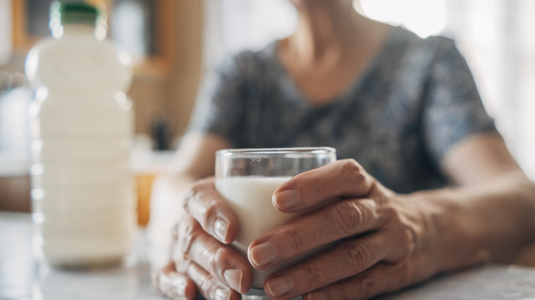 An older woman's hands holding a glass of kefir milk next to a bottle of kefir