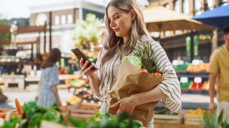 woman carrying bag of produce while looking at phone