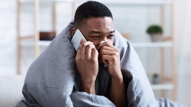 Sick man wrapped in blanket holding tissue speaking on cell phone