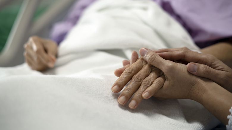 A younger woman's hand holds an older woman's hand on a hospital bed