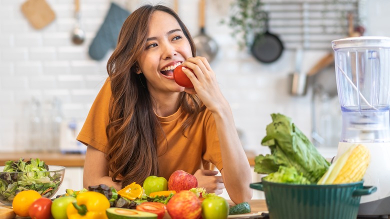 A young woman eats an apple while surrounded by fruits and vegetables