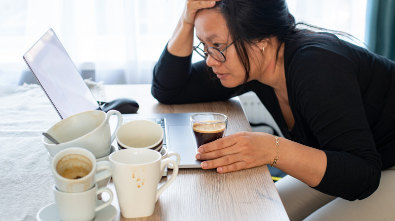 Stressed woman looking at computer screen surrounded by 8 cups of coffee