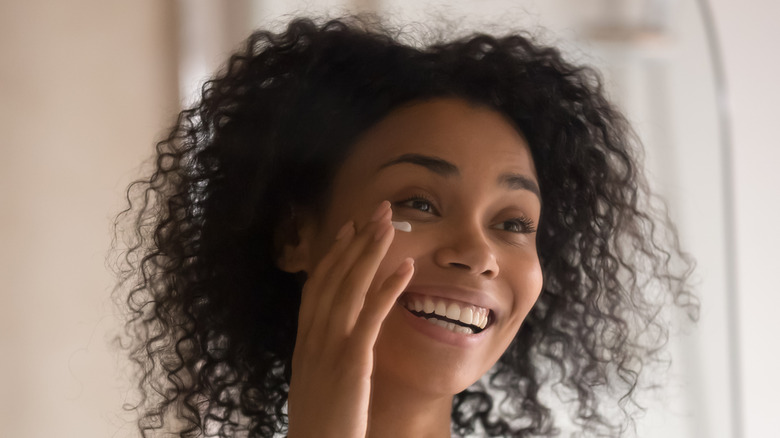 Smiling African American woman wrapped in towel after shower looks in mirror applying under eye cream