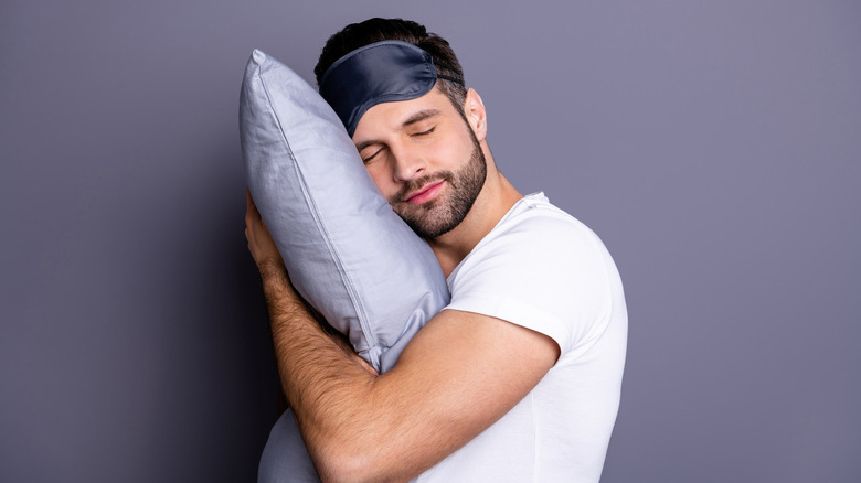 man hugging his pillow close with a sleep mask on his forehead 