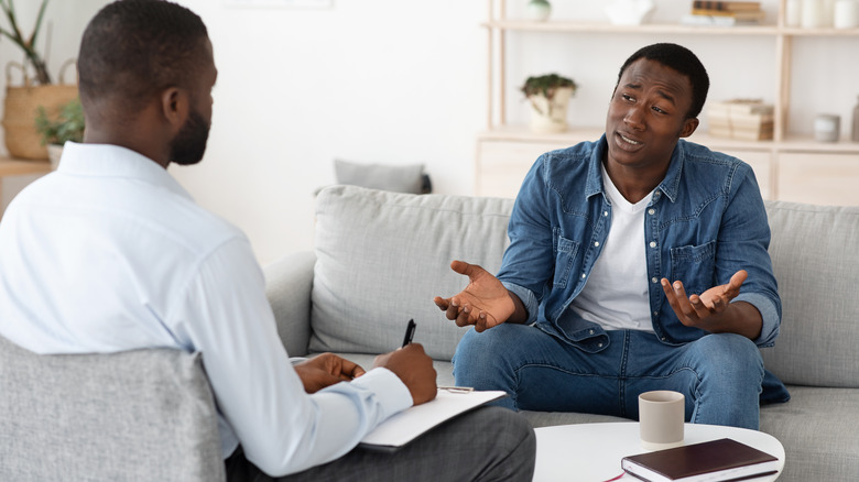 Young man sitting on couch talking to a therapist holding a pen and clipboard