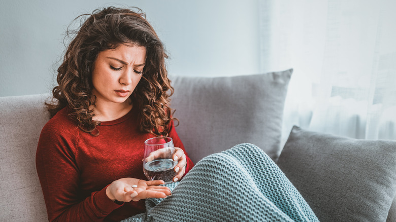 Woman sits on couch in pain with painkillers and a glass of water in hand