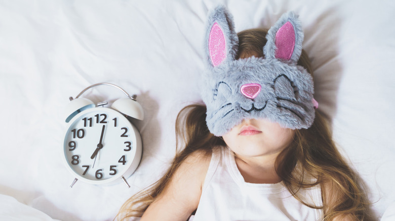 Girl with bunny sleep mask on, next to clock