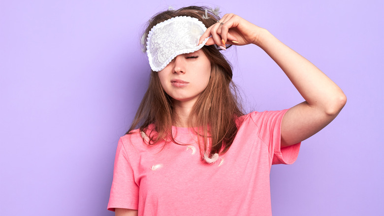 Exhausted woman in pink t-shirt slightly raises her white sleep mask, trying to open sleepy eyes