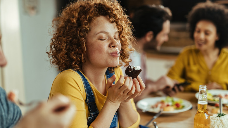 Woman with eyes closed enjoying taste of food while eating with friends at dining table.