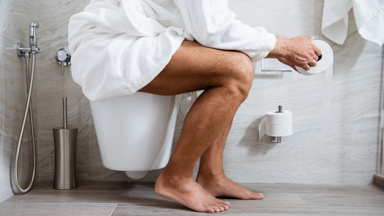 Close up of legs seated on toilet wearing a white robe and reaching for roll of toilet paper