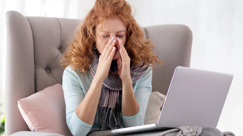 Woman sitting in chair with her laptop wearing a scarf and touching her nose