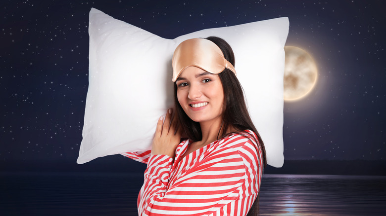 Woman in red striped pajamas and eye mask on forehead holding pillow with full moon in background