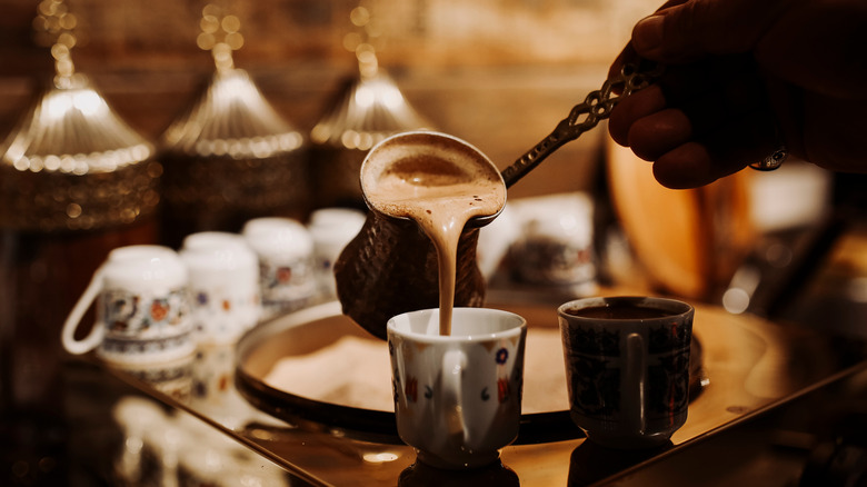 Hand pouring a mug of freshly boiled Greek coffee surrounded by china