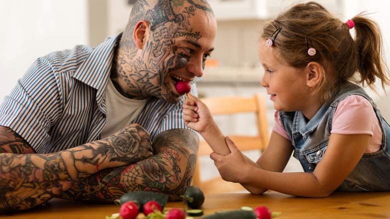 older man with tattoos being fed a radish by little girl