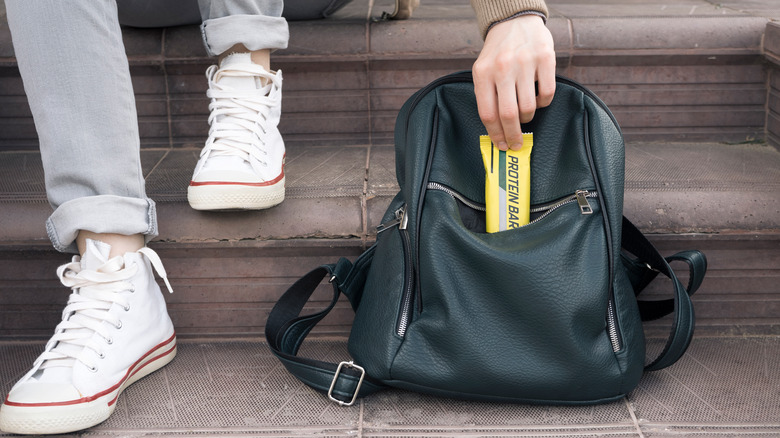 Close-up of a person crouched on steps pulling a protein bar out of their backpack. 