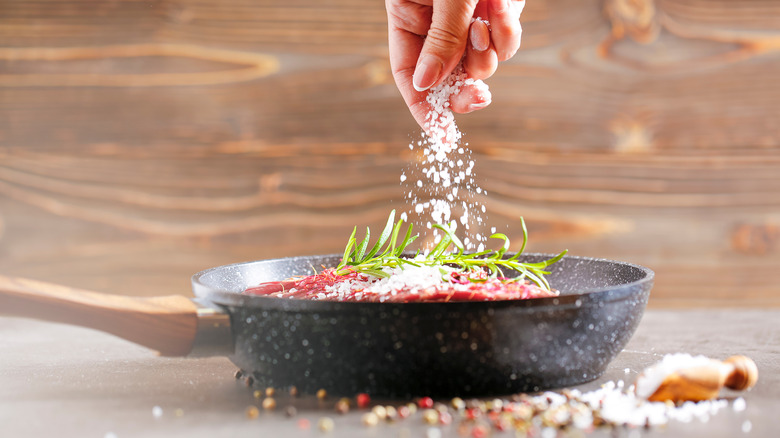 Someone adds salt to a pan of meat