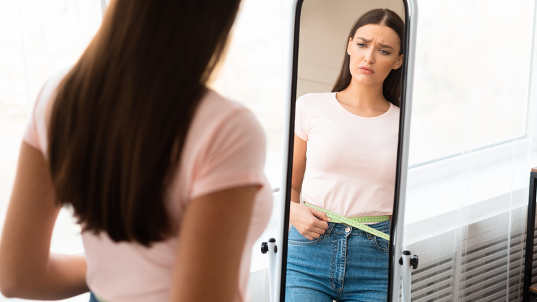 Woman holding tape measure around waist while looking in mirror