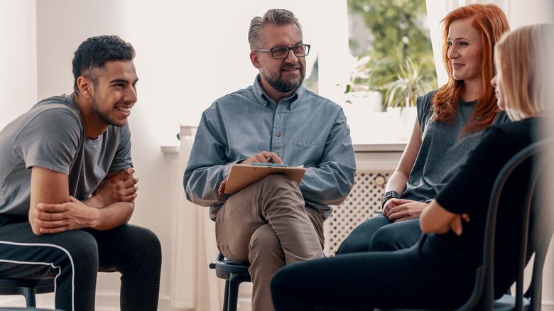 Man wearing glasses and holding a clipboard leading a group therapy session with 3 patients