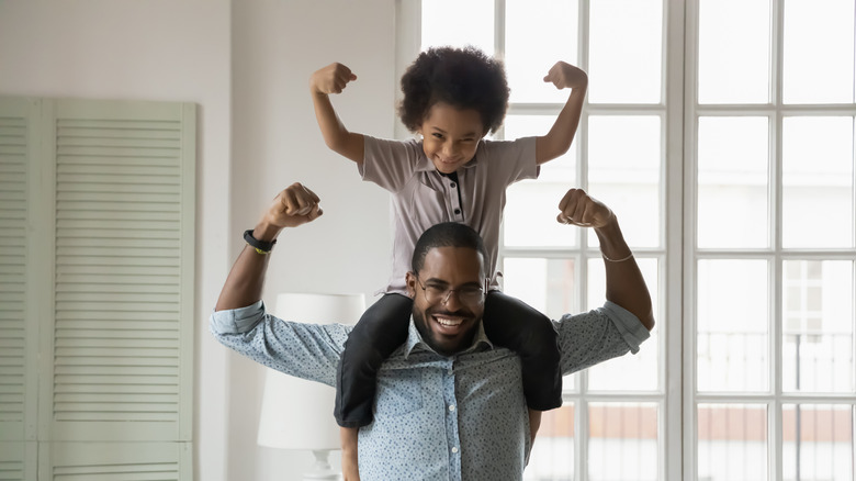 Small son sitting on dad's shoulders showing biceps.