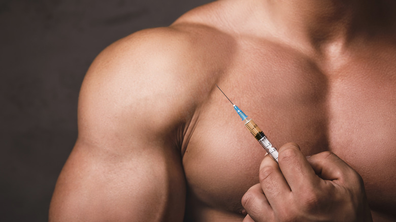 Close up of a muscular man's shoulder, pecks, and part of his hand as he holds up a syringe