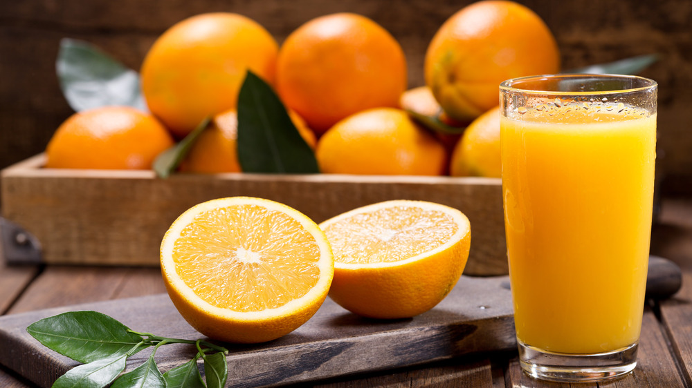 A glass of orange juice next to two halves of an orange and in front of a bunch of oranges