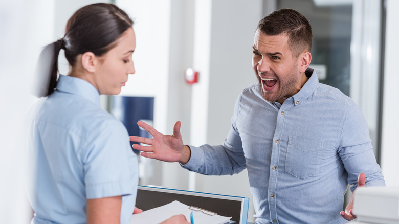 A man in a light blue shirt yelling at a nurse with a clipboard in a doctor's office