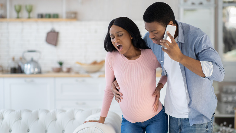 Pregnant woman experiencing a labor contraction while her partner has his arm around her and makes a phone call