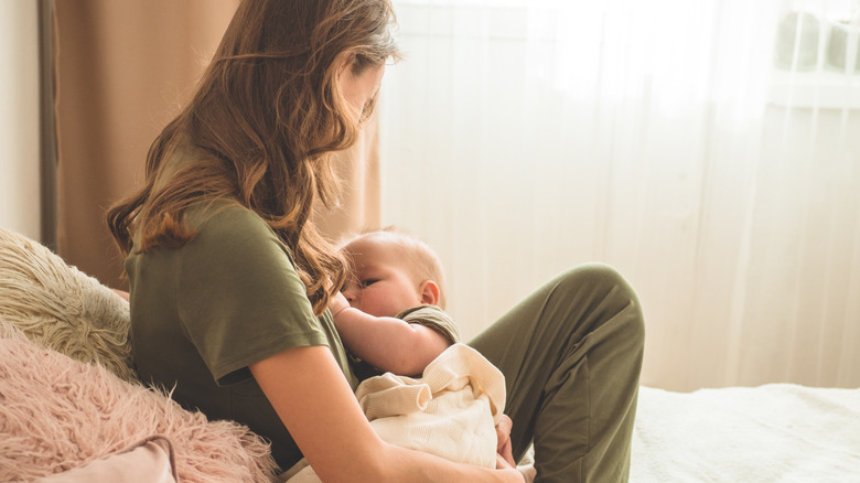 Mother breastfeeds her baby sitting up on bed