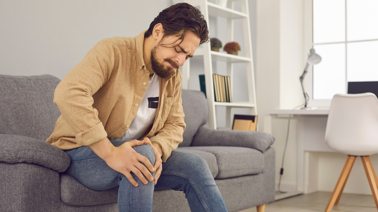 Man sitting on couch at home, grimacing while holding his knee
