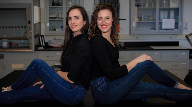 Erin Welsh and Erin Updyke of "This Podcast Will Kill You" sitting on a table in a lab