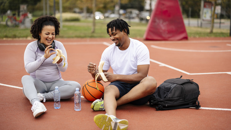 couple peeling bananas while sitting on a basketball court