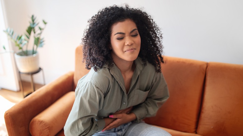 A woman holds her stomach in pain while sitting on her couch