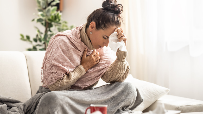 woman sitting on her couch wrapped in blanket and holding her forehead