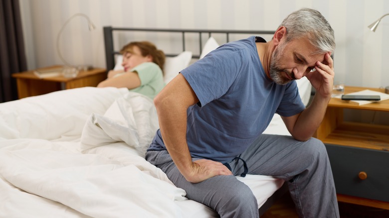 middle-aged man sitting on edge of bed while wife is sleeping