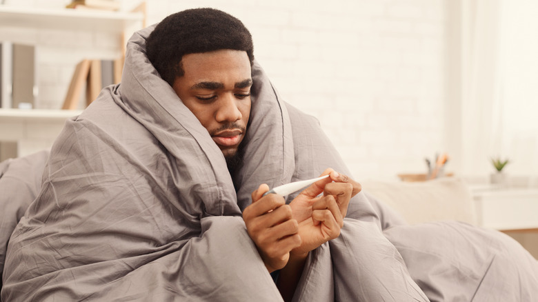 seated man wrapped in blanket looking at thermometer