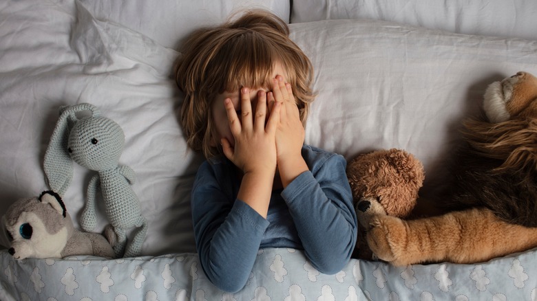 Child laying in bed covering face with hands