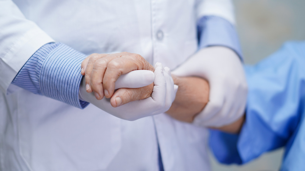 A person in a white medical coat and gloves holds the hands of another person wearing a medical gown