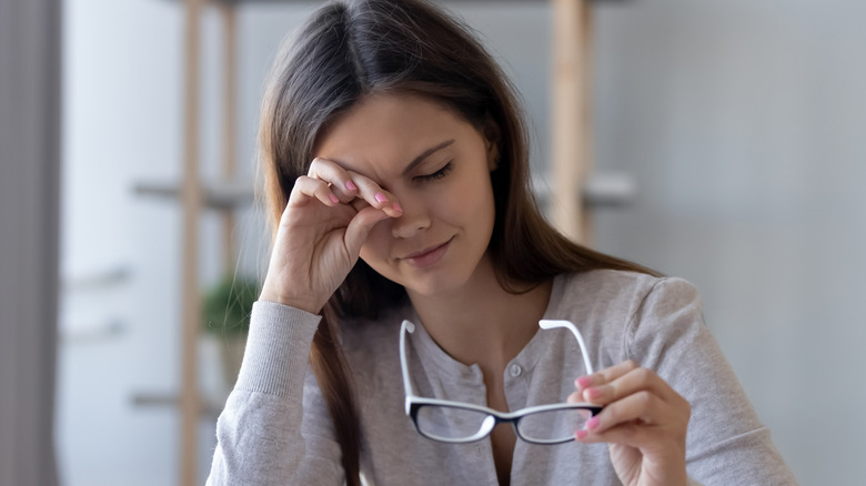 Young woman holding a pair of eyeglasses and rubbing her eye in discomfort