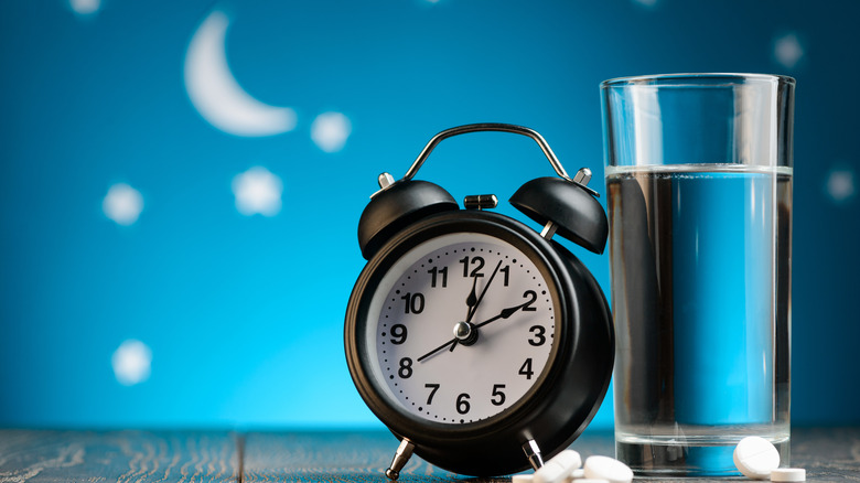 a clock with some nighttime pills and a glass of water with a crescent moon in the background