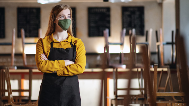 Masked restaurant worker stands in an empty restaurant wearing a face mask with arms crossed