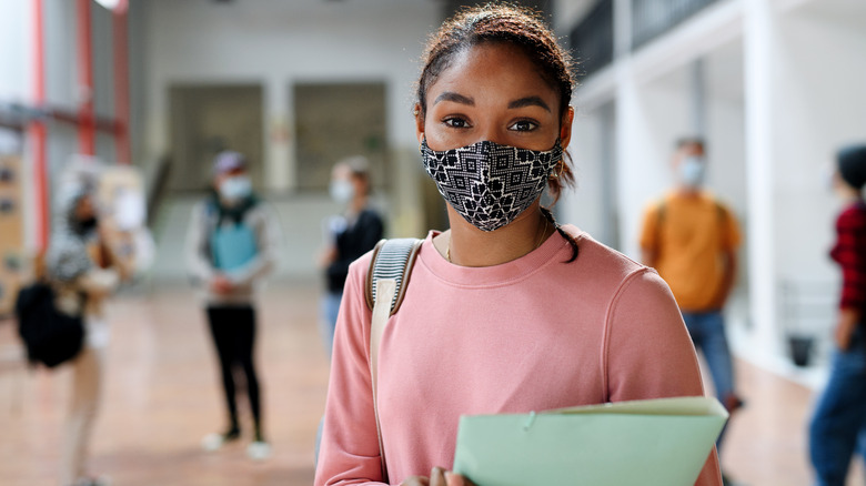 Student wearing a face mask and holding a binder standing in a school hallway amongst other students