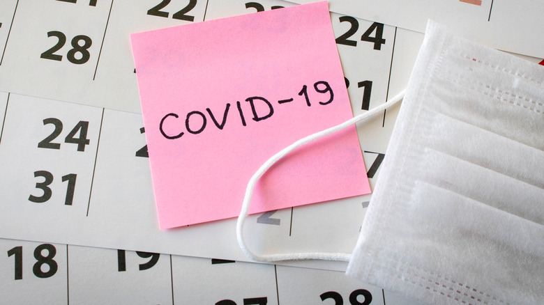 Pink sticky note reading "COVID-19" stuck on a calendar next to a surgical face mask