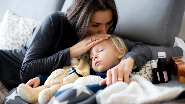 Sick toddler sleeps on the couch with their mother kissing them on the head. Surrounded by thermometer and bottles of medicine