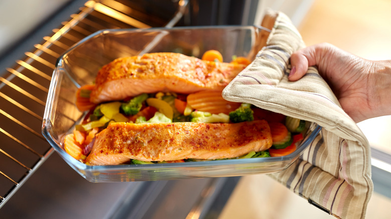Hand placing a glass pan of salmon and veggies into oven