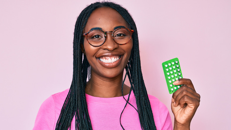 Woman smiling at camera holding birth control pack against pink background