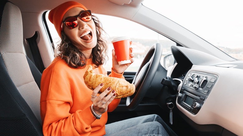 Woman eating coffee and a croissant in the car and smiling  