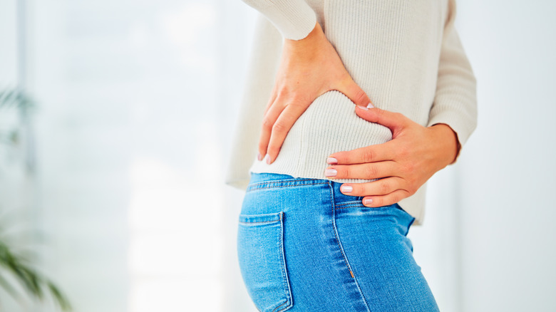 Person wearing white top and blue jeans holding hip with two hands as if in pain