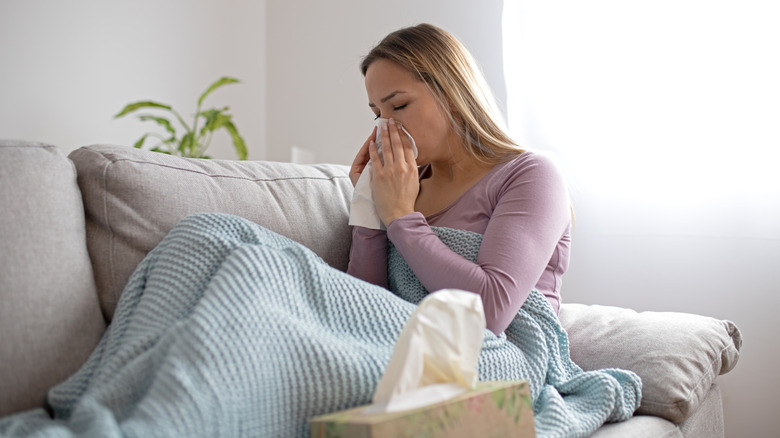 A woman blowing her nose while seated under a blanket on her couch